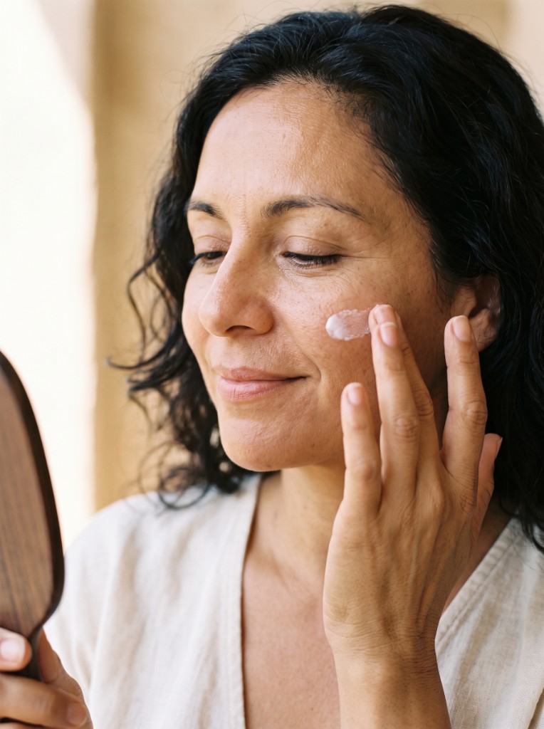 Woman applying cream to her cheek while looking into a wooden hand mirror