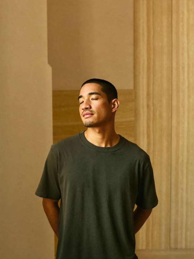 Model in a dark olive T-shirt standing with eyes closed in a quiet stone room