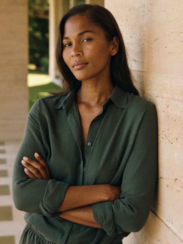 Model in a forest-green shirt with arms crossed, leaning on a stone wall