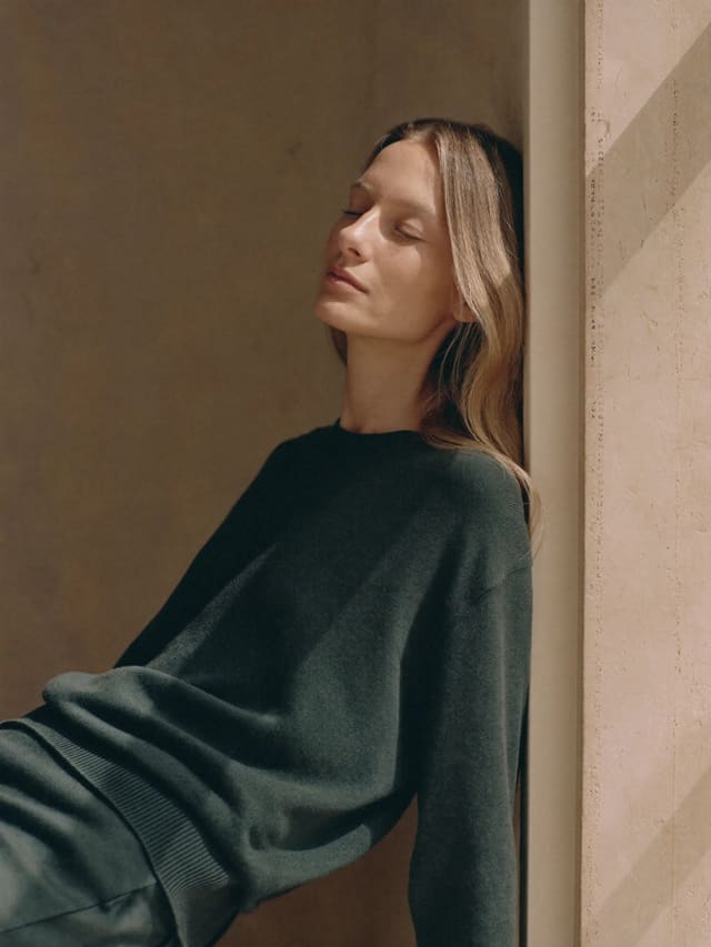 Model in a forest-green sweater leaning into a sunlit doorway, eyes closed