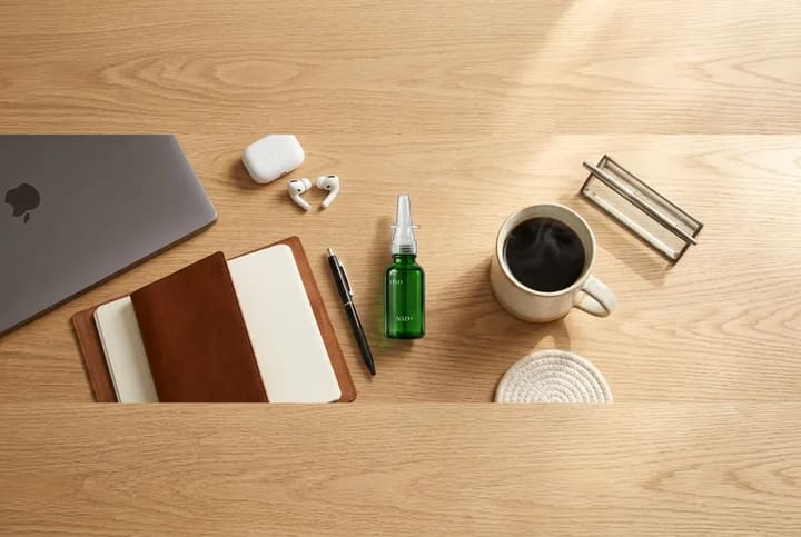 A green NAD+ nasal spray bottle on a wooden desk beside a notebook, pen, earbuds and a cup of coffee