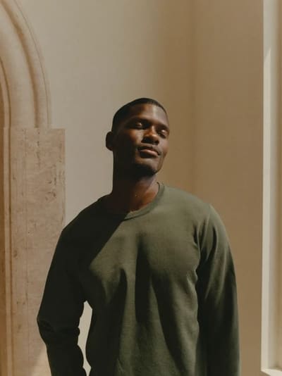 Model in an olive sweater standing in soft light beside a stone arch, eyes closed